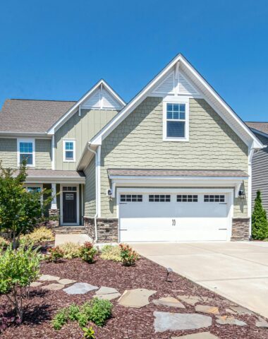 Charming suburban house with green facade, garage, and landscaped yard on a clear day.